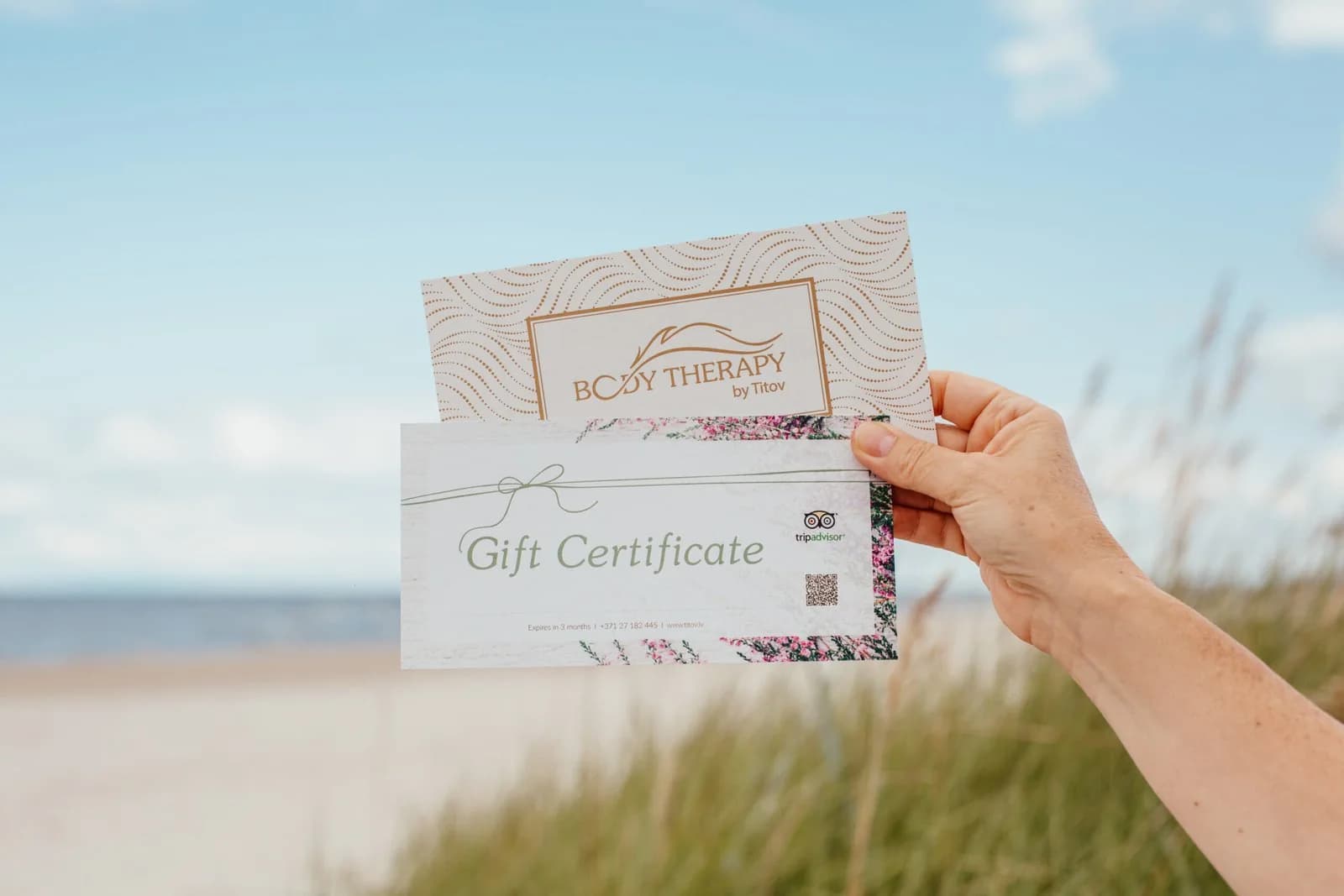Coach and massage therapist Aleksejs Titovs holding Therapy by Titov gift certificates by the seaside in Riga, with blue sky and beach in the background.