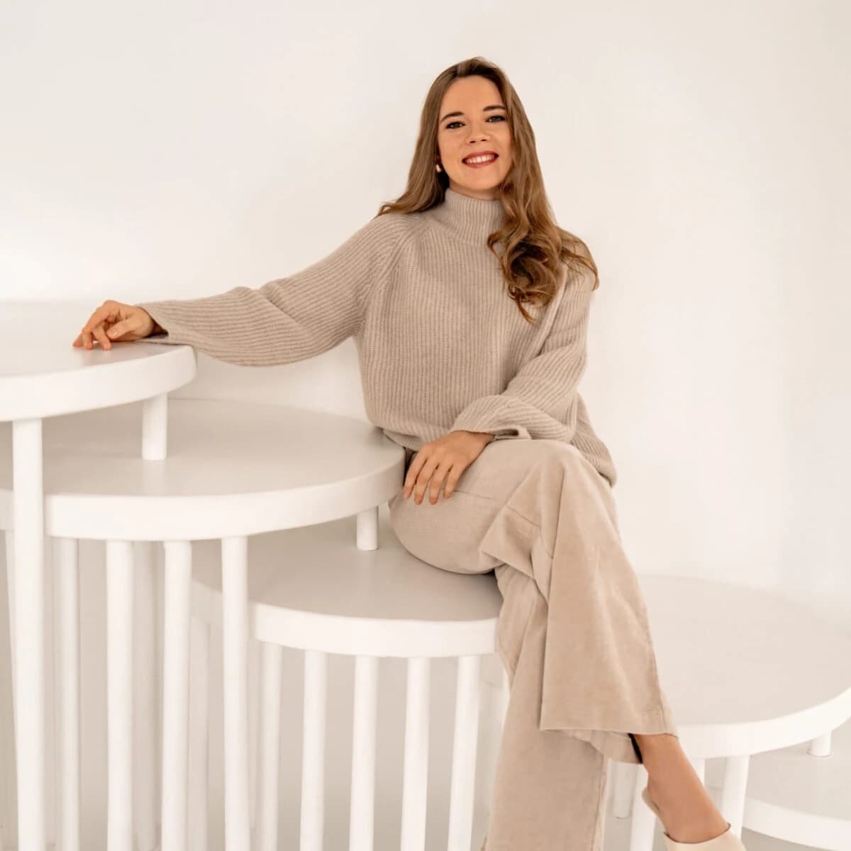 Therapy by Titov terapeite Darja Titova gaišā studijā sēž uz baltām pakāpēm, simbolizējot unikālu un detaļām pievērstu darbu ar katru cilvēku.